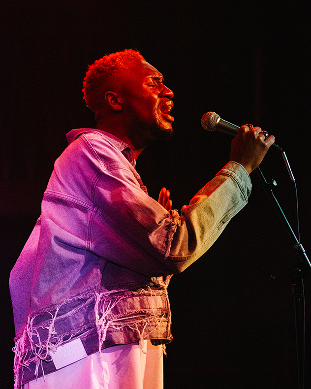 A man singing on stage lit in a pink and purple hue.